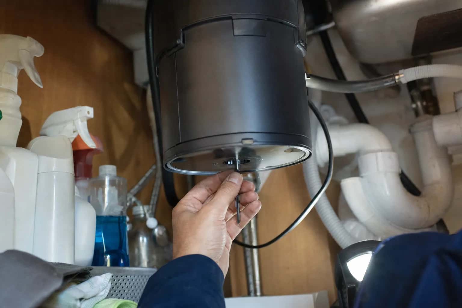 A hand reaching under a kitchen sink to the bottom of a garbage disposal