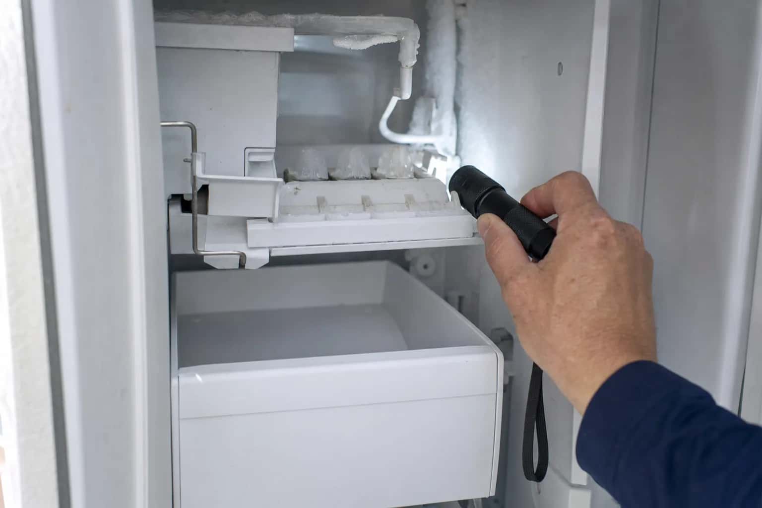 A flashlight aimed at an empty ice maker and ice bin inside a freezer