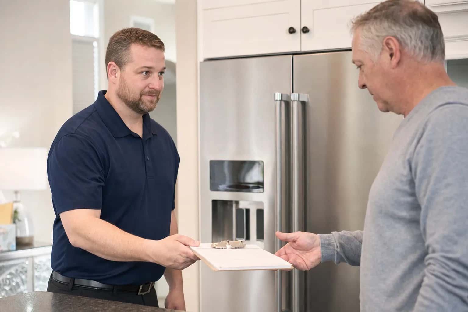 A man in a navy polo handing a written estimate to a homeowner beside a stainless refrigerator